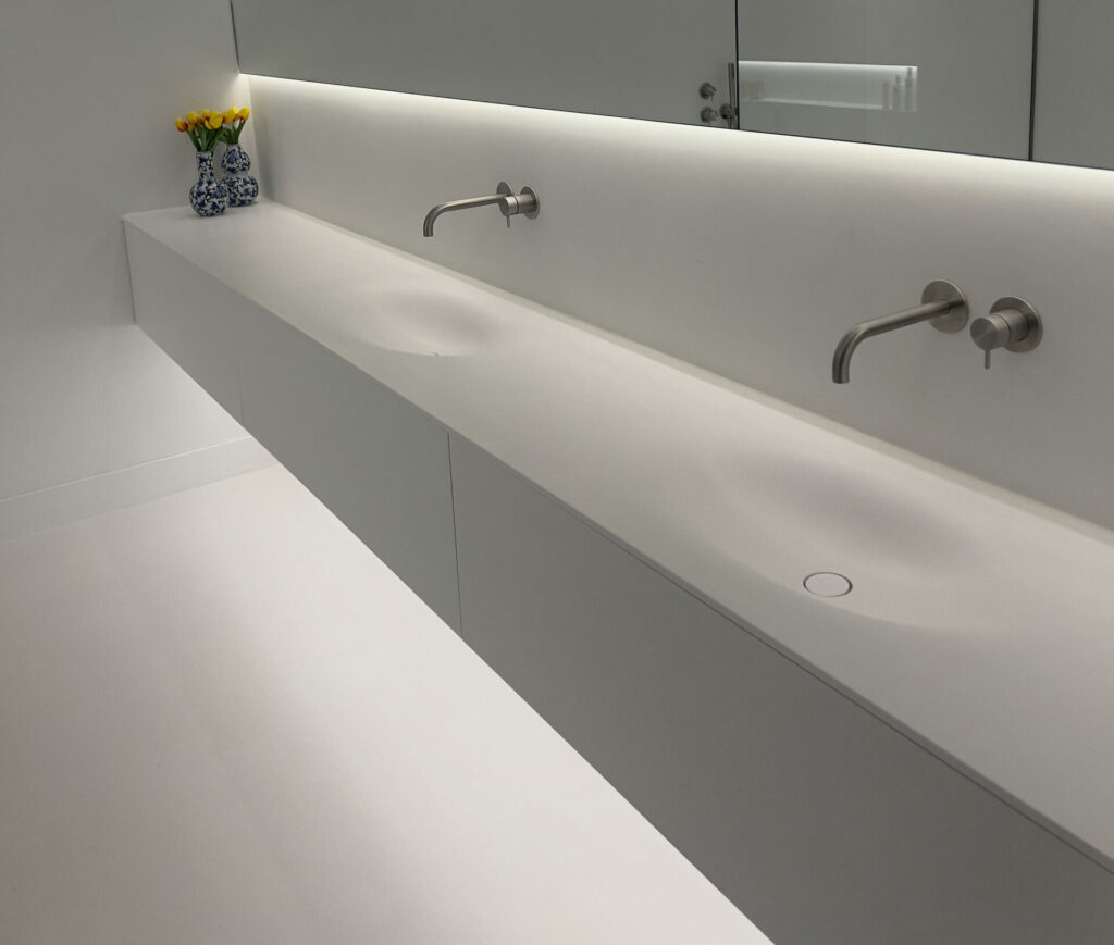 Wandbrede witte wastafel met een langwerpige, naadloos verzonken wasbak en twee wandkranen, onder een verlichte spiegel